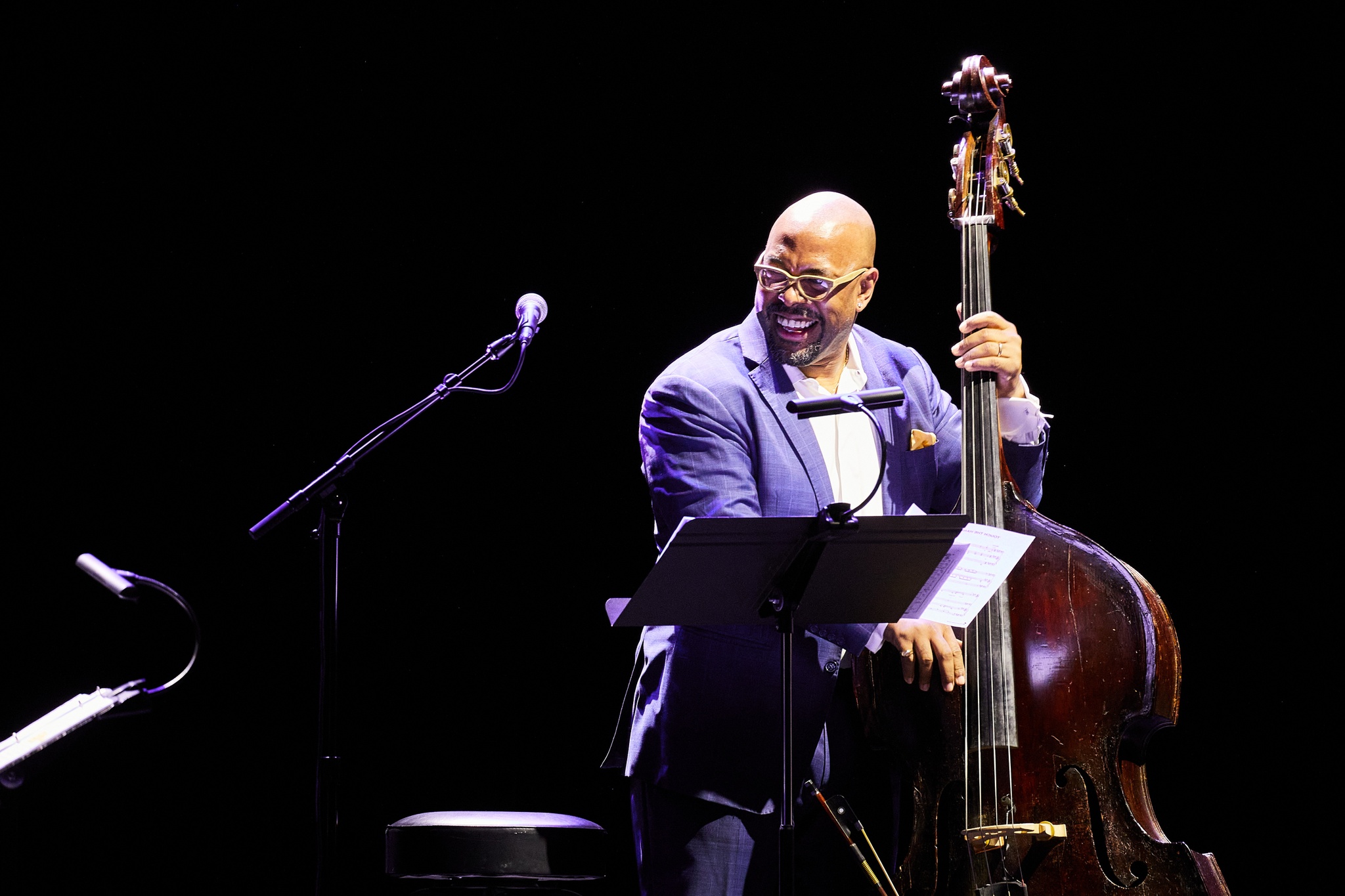 A bassist holding his instrument and laughing while on stage performing. He is a Black man with a bald head and he wears a blue jacket.