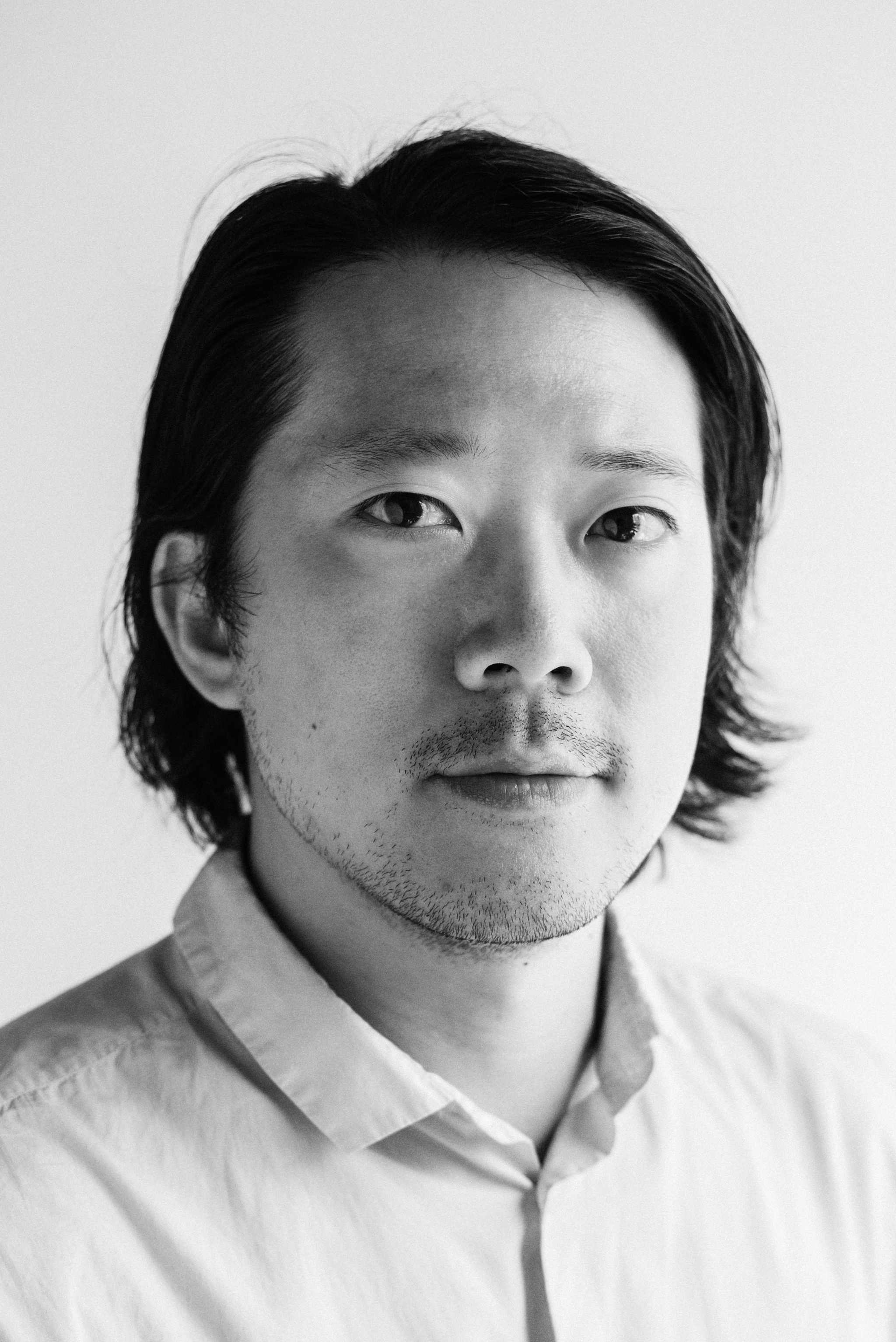 A black and white headshot of Kenneth Tam, an Asian American man who poses against a blank background. Kenneth has hair parted down the side of his head that he tucks behind his right ear. He looks pensively at us. 