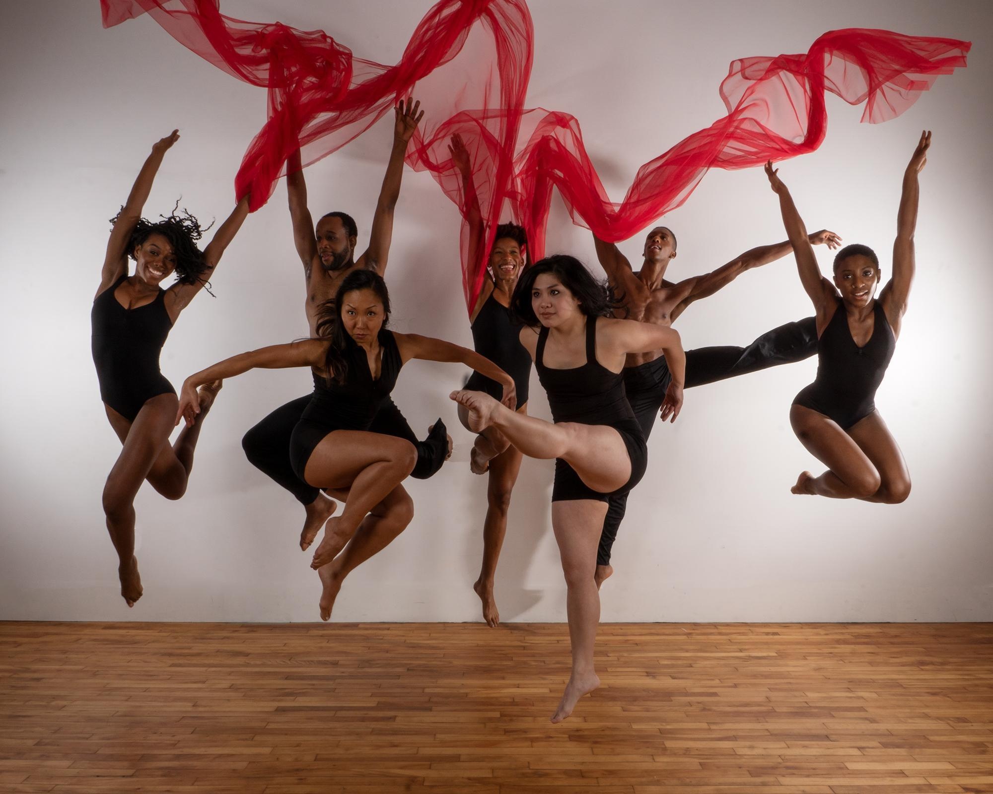 Members of the Rod Rodgers Dance Company leaping and jumping in the air. They are dressed in leotards and dance gear and stretch red gauzy fabric above their heads.