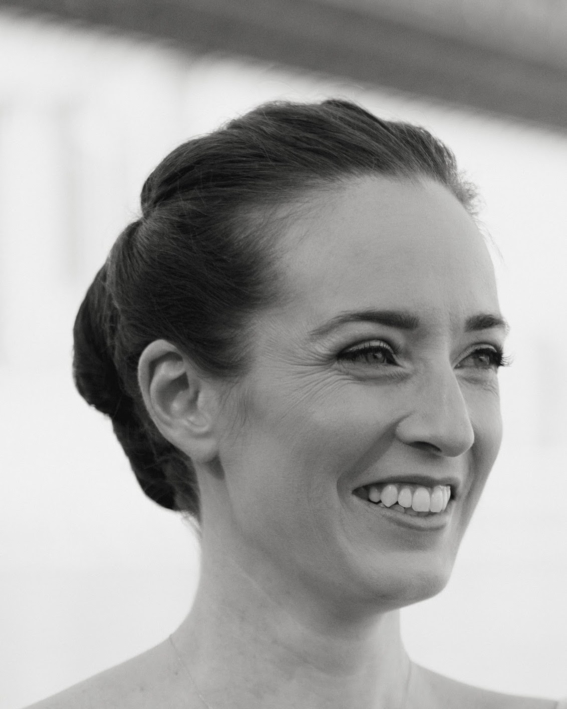 A portrait of dancer Julie McMillan Castellano, who smiles with her eyes squinting and her lips open revealing the top row of her teeth. She wears her hair pulled back in a low bun. 