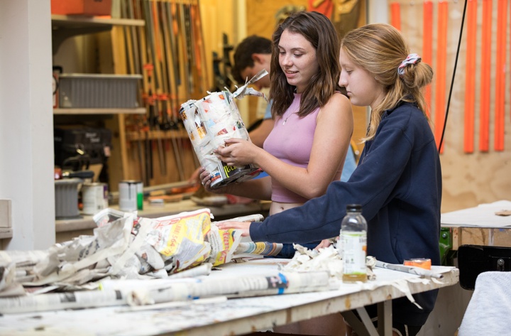 Two people work on a papier mache project at a worktable in a shop space.