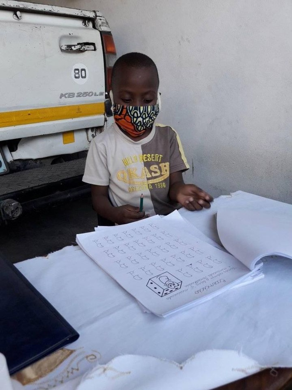 A color photograph shows young dark-skinned boy wearing a mask, studying, and reading educational materials.