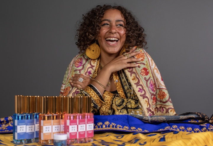 Portrait photo of Noor Bekhiet, wearing a vibrantly patterned, brightly colored garment and large round yellow earrings. In front of Noor is a bright blue and gold fabric, with three rows of three bottles in front. The bottles all have gold tops; the three on the left are blue, the three in the middle are orange, and the three on right are bright pink.
