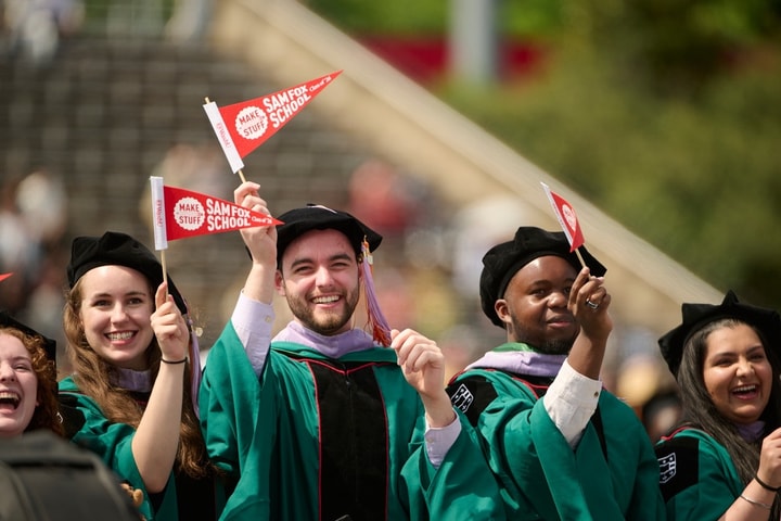 A group of students wearing regalia stand and wave their Sam Fox School pennants.
