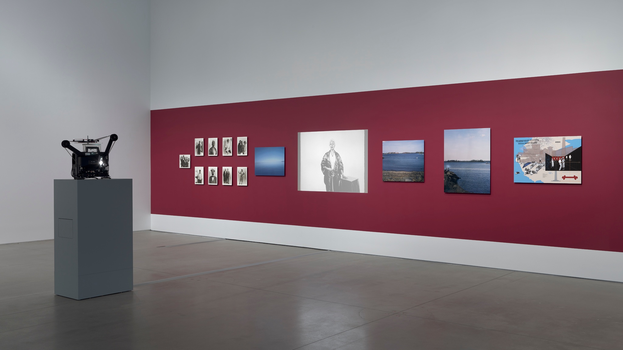 A film projector sits on a plinth at a distance from a red gallery wall with photos of different sizes hung on it