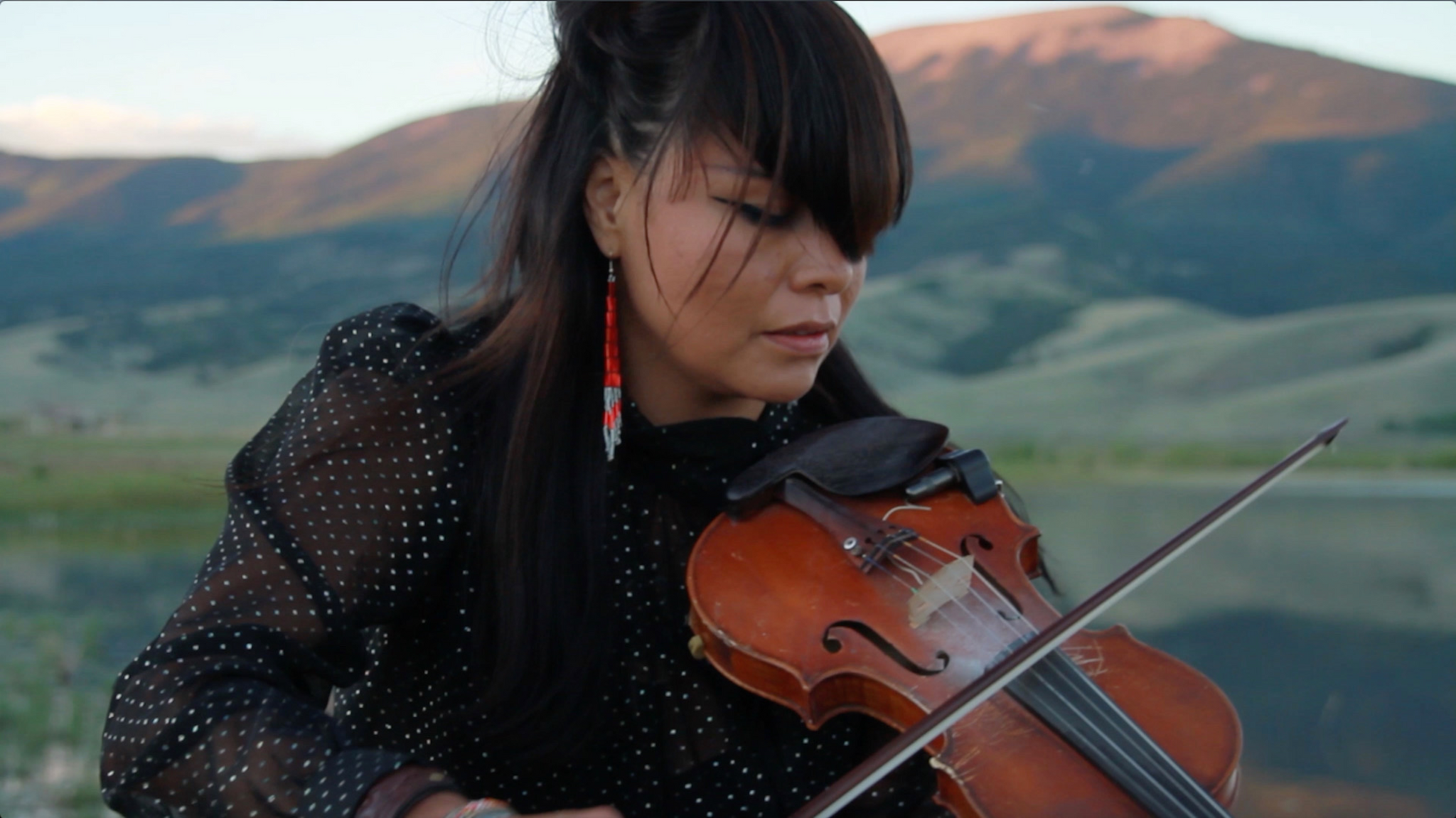 A portrait of Laura Ortman playing a violin outside. She has long bangs that fall over her face and wears a black blouse with puffy, sheer sleaves. Behind her a mountain rises in the distance. The top of the mountain is illuminated by sunlight.