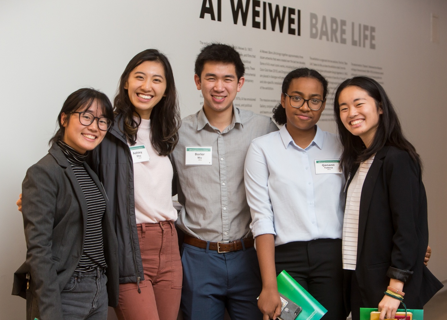 Five people pose in front of a white wall with vinyl lettering reading "Ai Weiwei Bare Life."