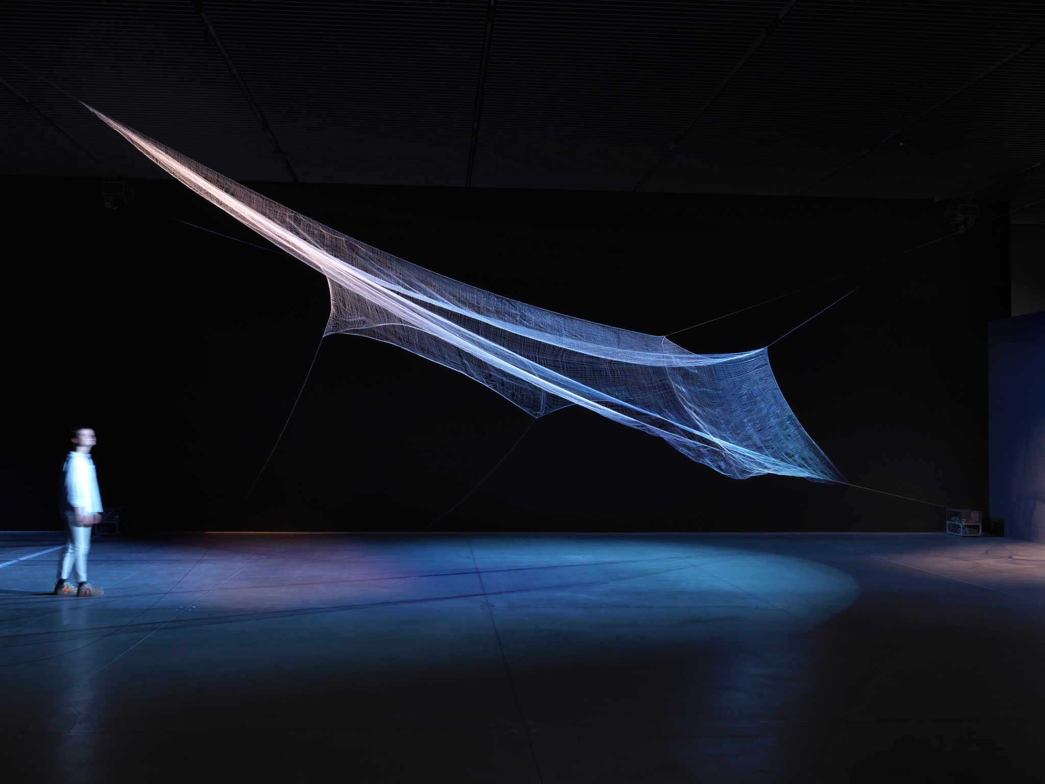 A fabric sculpture stretched elegantly in an upward slope toward a solitary viewer in a darkened gallery. The sculpture is lit in a soft blue and purple light.