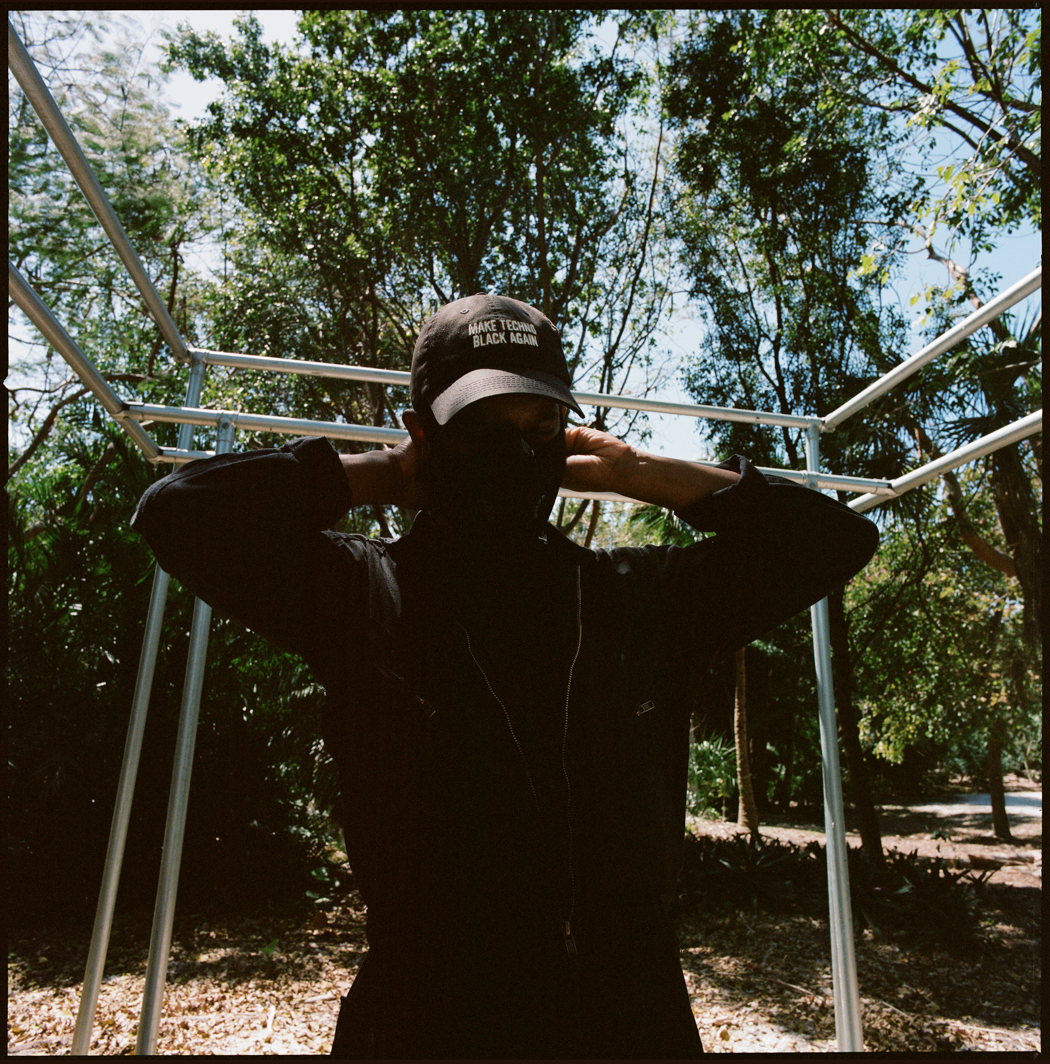 A photo of DeForrest Brown Jr. against a backdrop of trees. Brown's face and torso are in shadow and his hands are behind his head adjusting a face mask. 