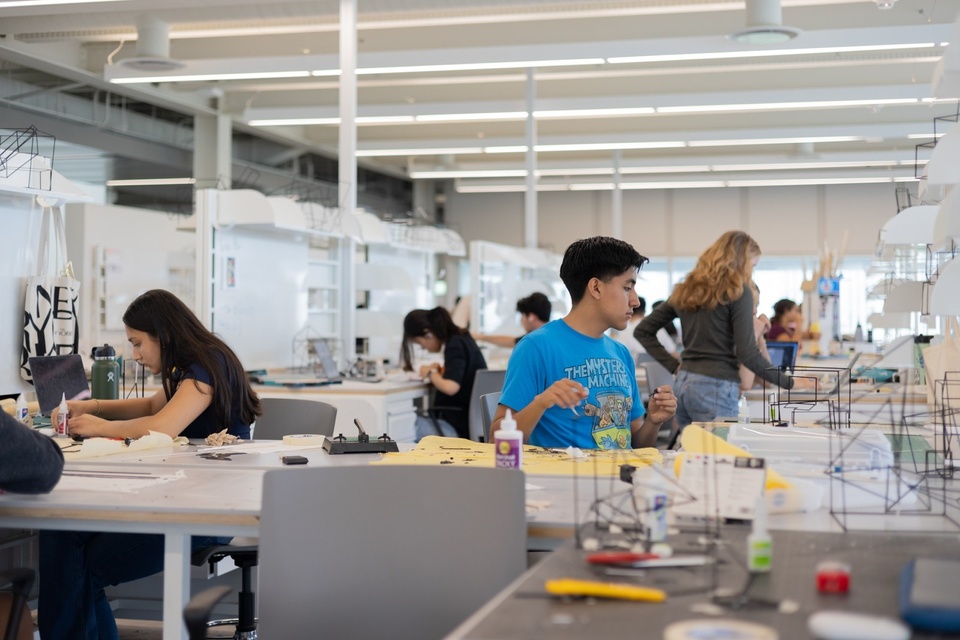 Several students in a large studio sit at tables working on architectural models.