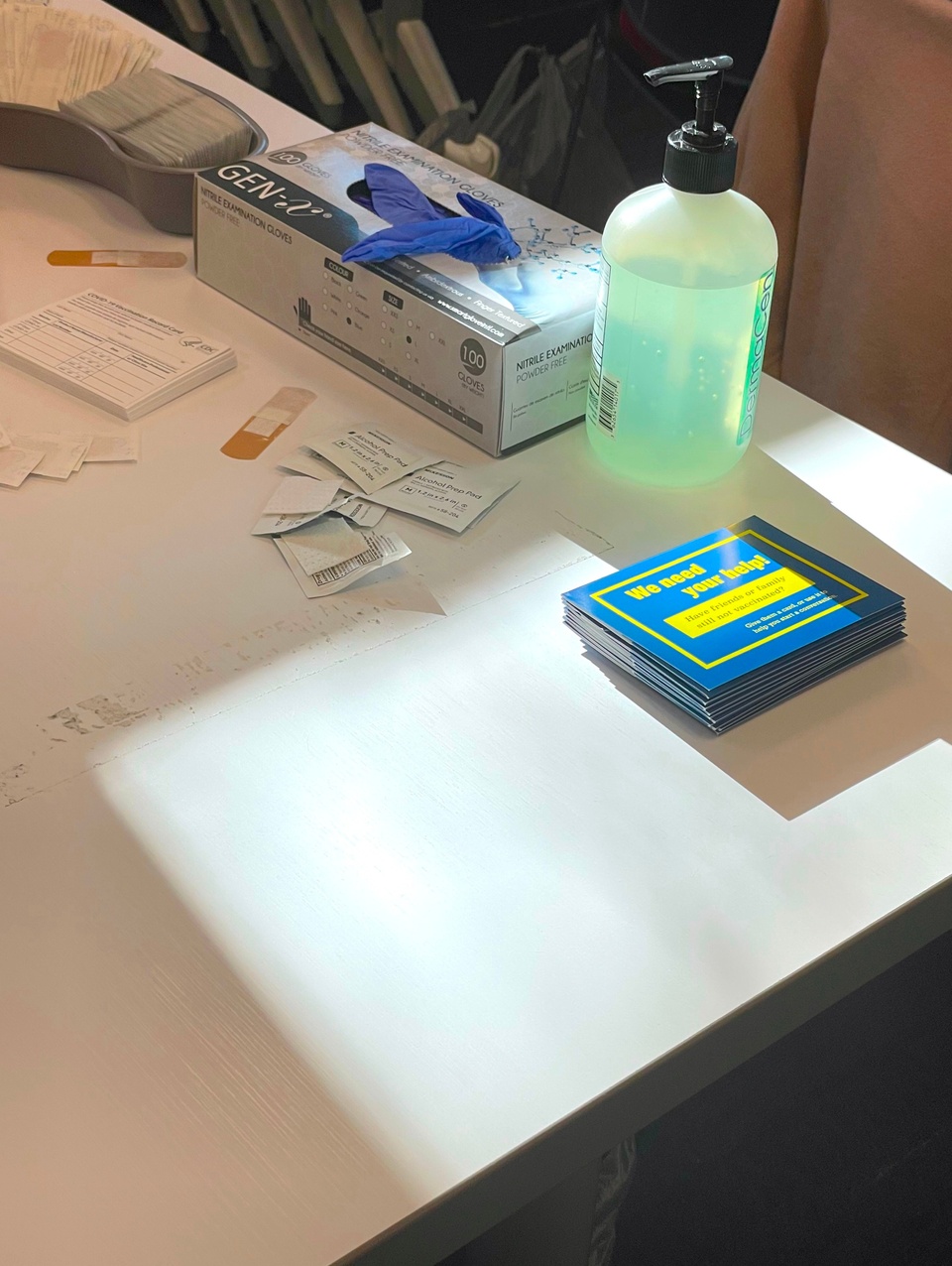 A stack of blue card holders on a desk at a vaccination event. Surrounded by a bottle of hand sanitizer, bandaids, a box of gloves and miscellaneous papers.