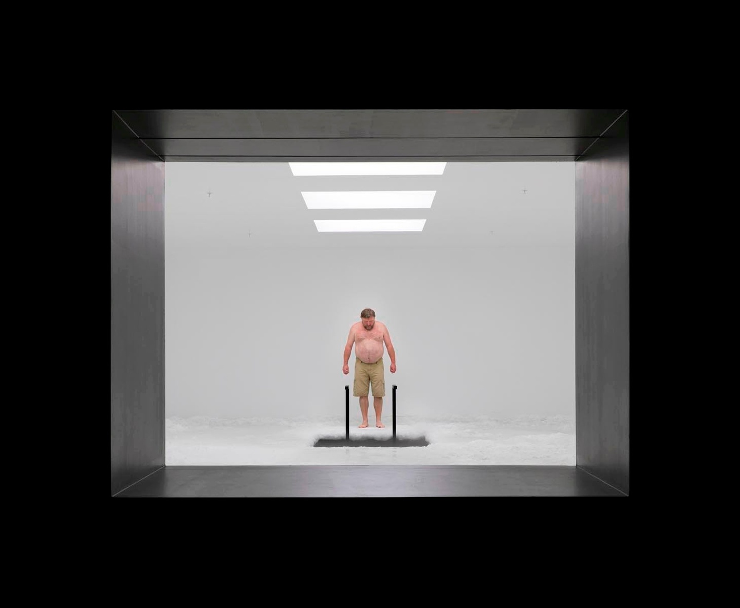 A black frame around a view into threshehold of a white room with three rectangular, overhead lights. A shirtless individual is looking down at a rectangular opening in the floor, with what might be a black ladder leading into it.