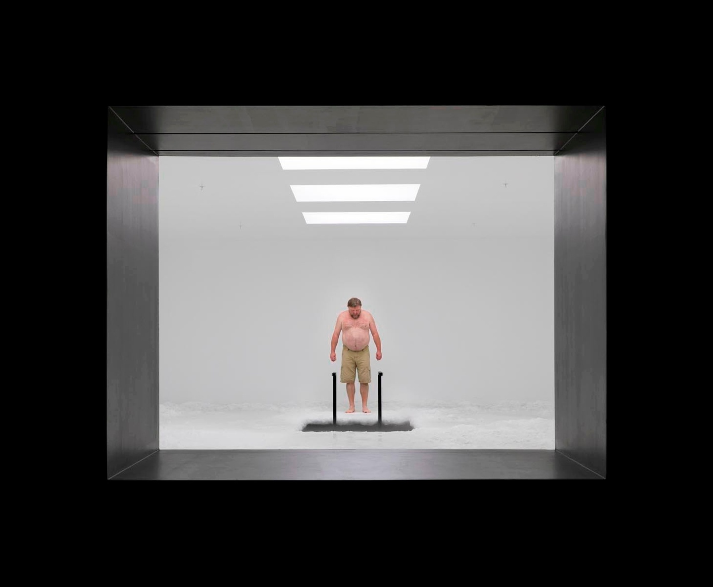 A black frame around a view into threshehold of a white room with three rectangular, overhead lights. A shirtless individual is looking down at a rectangular opening in the floor, with what might be a black ladder leading into it.