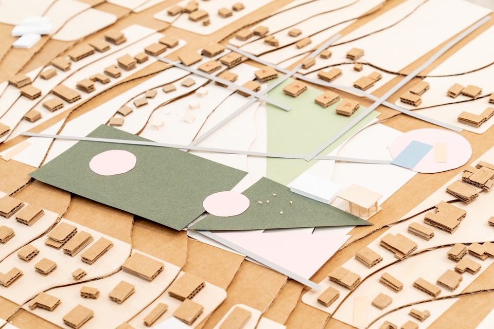 Overhead shot of a city plan model showing topography, buildings, and a central park area made out of cardboard and olive, pink, and tan paper.