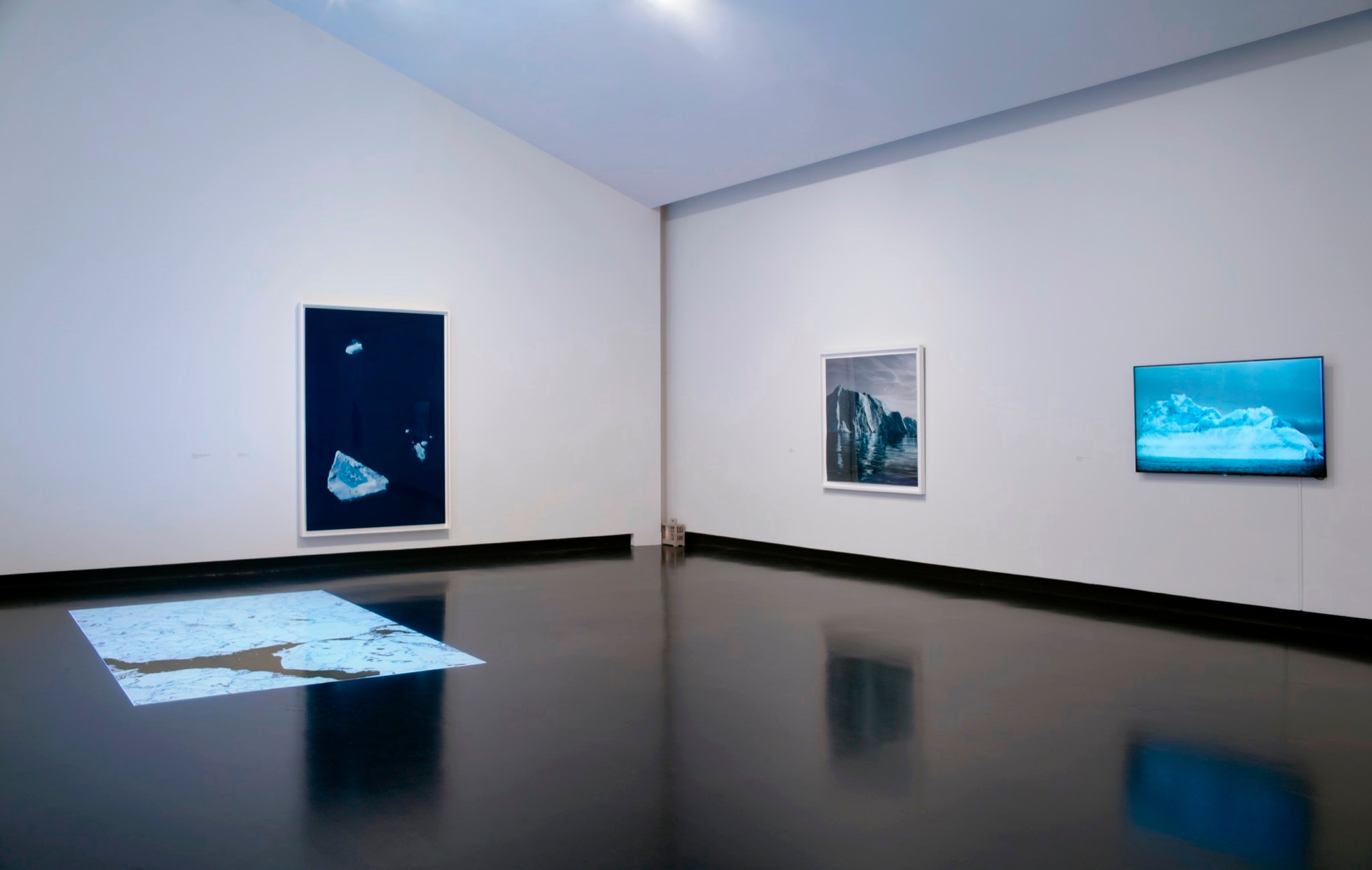 Displayed in a museum gallery with white walls and a black floor are four 2-D artworks featuring dark blue, light blue, and white.