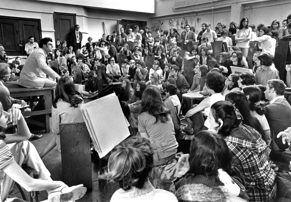 Black and white photo of a packed drawing studio. A person on a dais is speaking, dozens of others are gathered around listening, sitting on drawing horses.