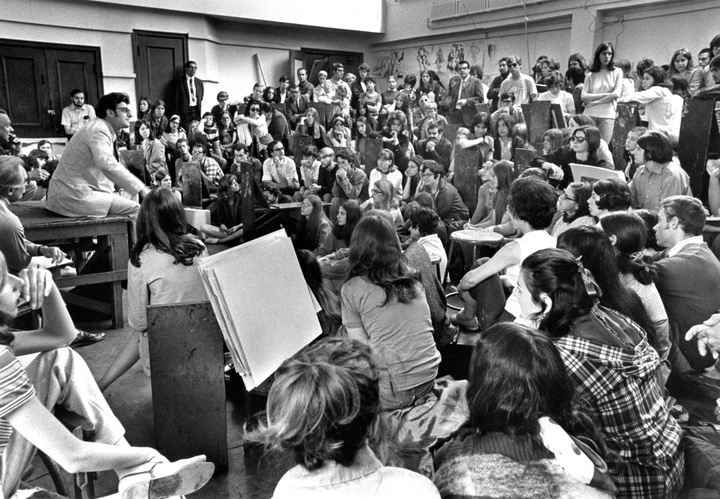 Black and white photo of a packed drawing studio. A person on a dais is speaking, dozens of others are gathered around listening, sitting on drawing horses.