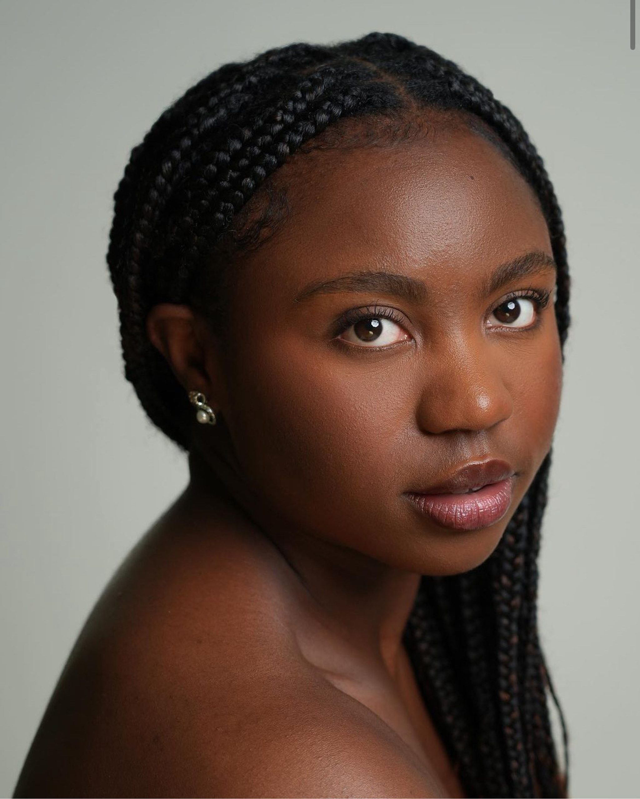 A headshot of dancer Libiya Gray, a Black woman who poses looking at us over one bare shoulder. Her braids are pulled around her head to hang down over her left shoulder. She looks directly at us expectantly. 