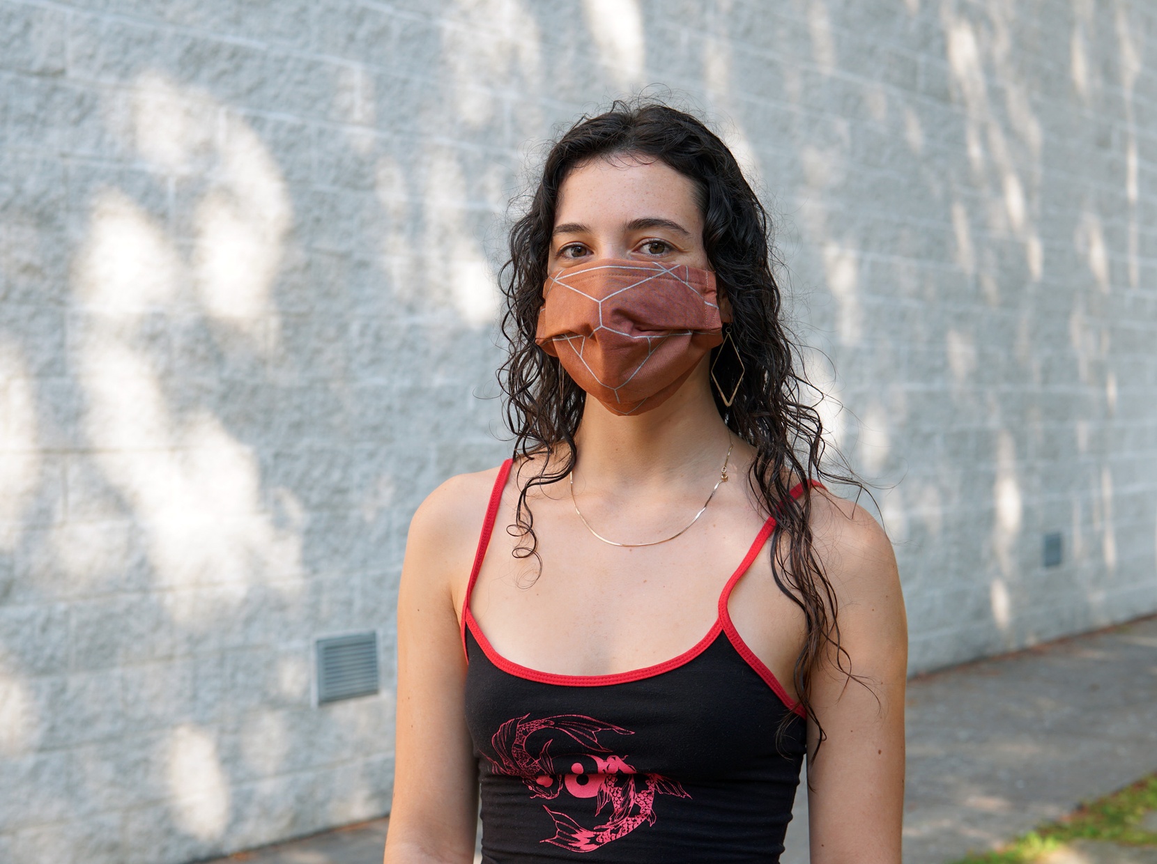 A light-skinned, young woman with dark hair wearing a reddish-orange mask with gray, geometric patterns printed on it.