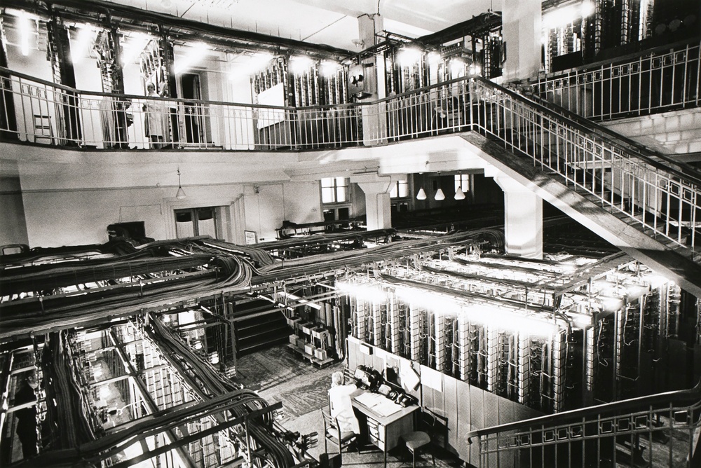 Black and white photograph of a two-story room filled with complex technical equipment containing lots of wires and lights.
