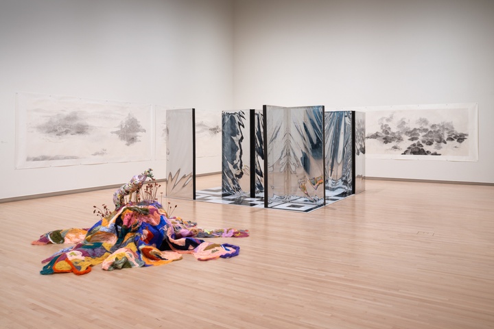 Overview shot of gallery showing a soft colorful felt sculpture on the floor and construction with vertical mirrored panels.