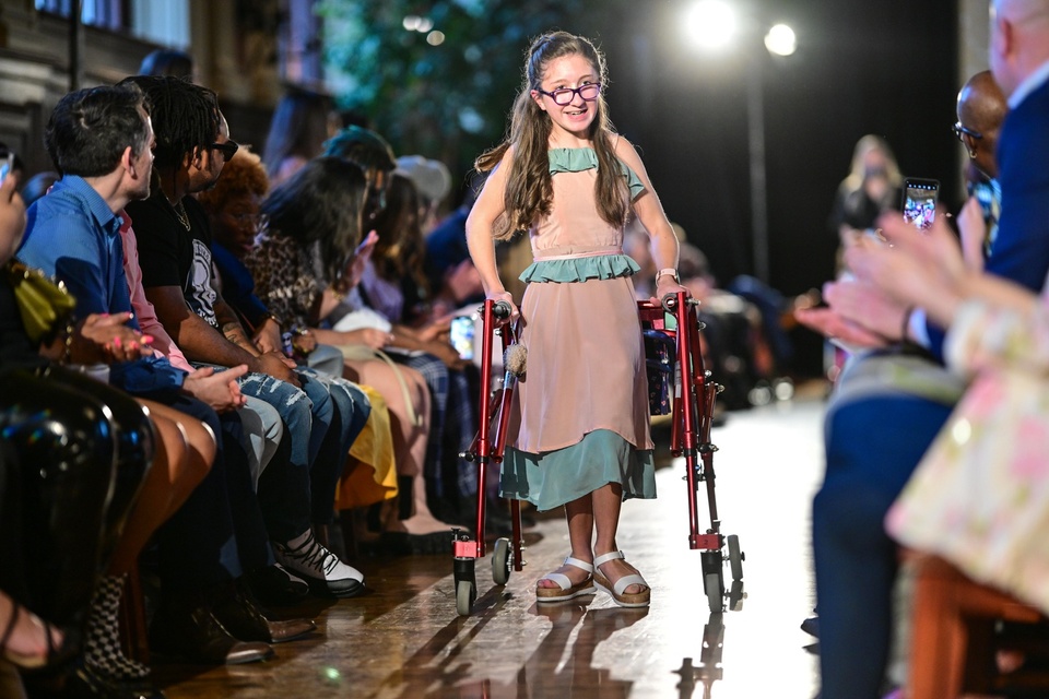 A model walks down the runway with the aid of a wheeled walker wearing a pink dress with teal ruffles