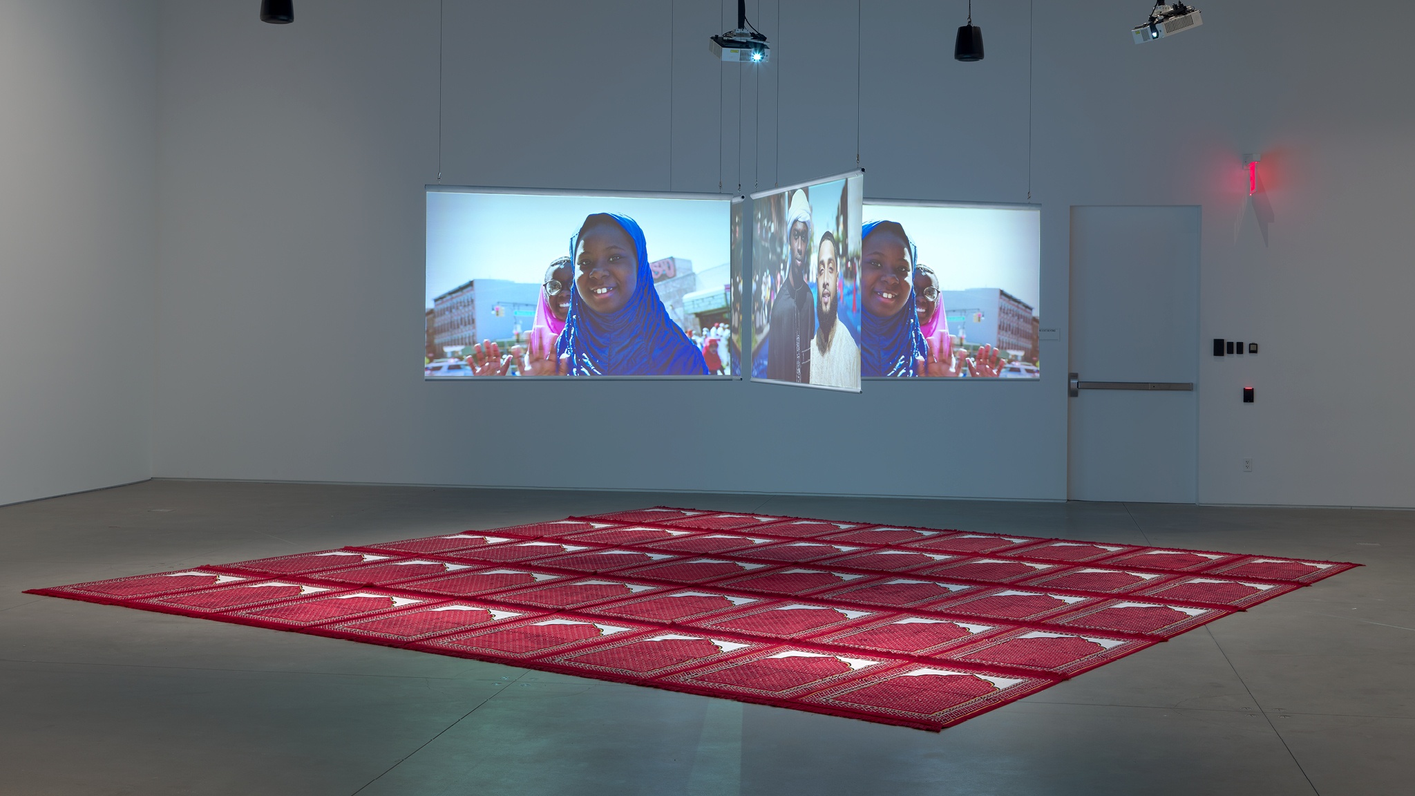 Three video screens suspended in an intersecting formation above a floor covered with red prayer mats placed side by side to form one large square. The screens show a young girl in a headscarf smiling at the camera.