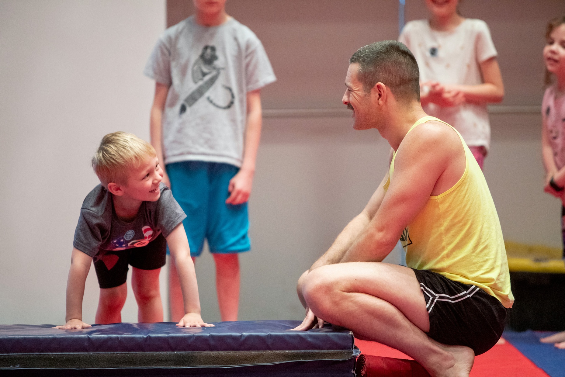 A little, white boy crouched on a blue mat and a male dancer smile at each other during a movement workshop.