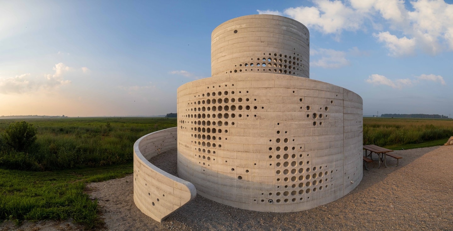A two-tiered cylindrical light-concrete structure featuring patterned circular cutouts and a curved spiral ramp, set in an open grassy field under a bright, softly clouded sky.