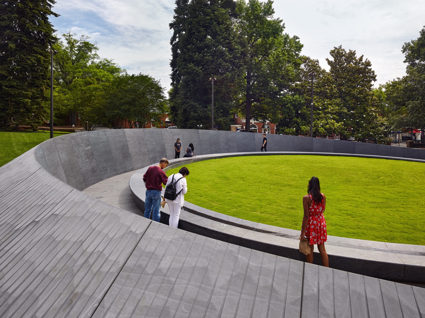 Photo showing a portion of the gray, curved memorial wall, with people walking inside it; the memorial surrounds a ovular patch of grass.