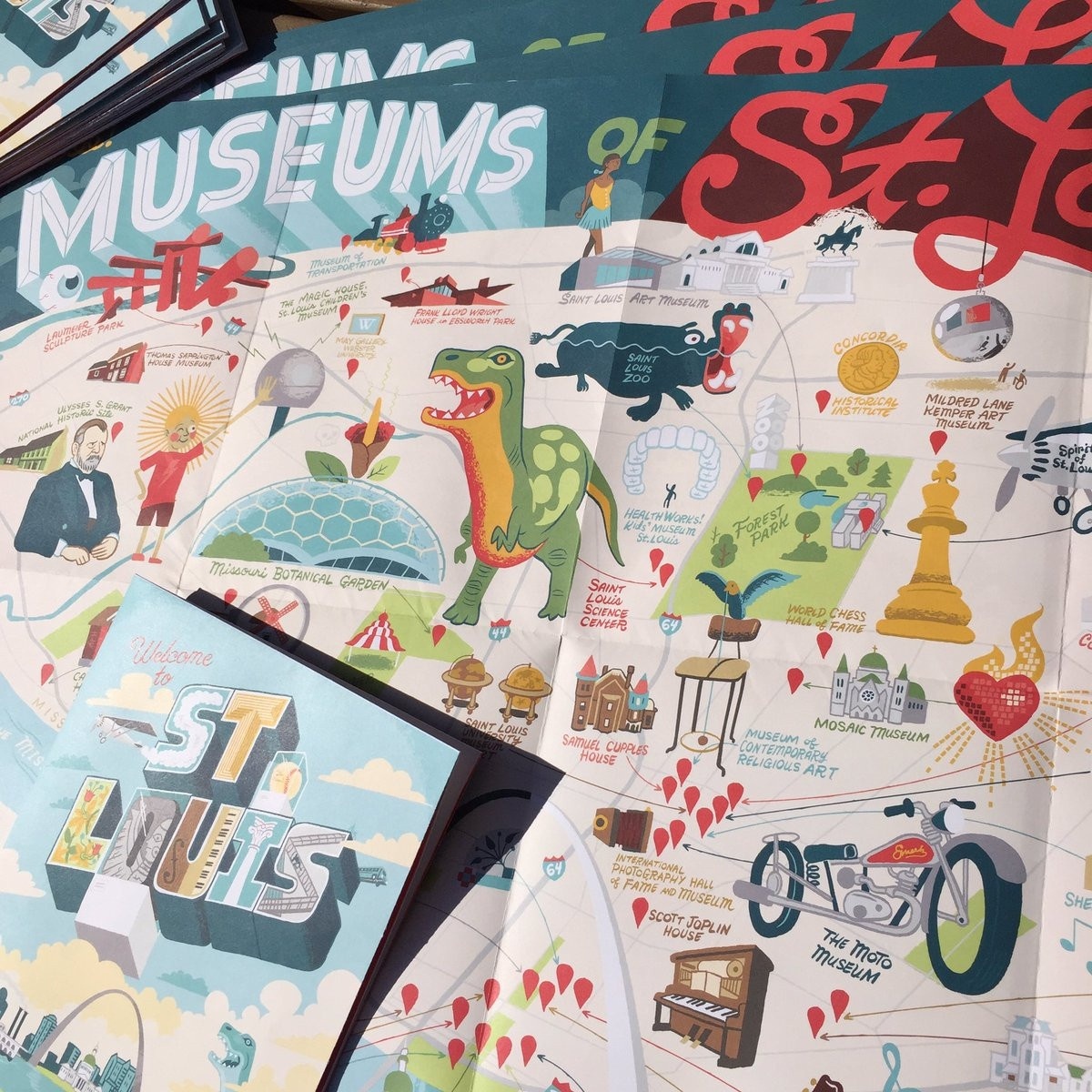 A brochure/poster folded out: the hand lettered title reads, "MUSEUMS OF St. Louis" and frames an intricate illustration of an assortment of various subjects, each a focus of a museum. The drawings are set on a light cream background that's a map, with red pins marking the location of each museum.