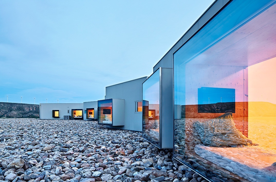 Photo of a contemporary, single-story hotel with gray, smooth walls interspersed with bumped out, square-shaped windows, the closest to the righthand of the frame revealing warm light and a bed. The hotel is built atop a bed of rocks.