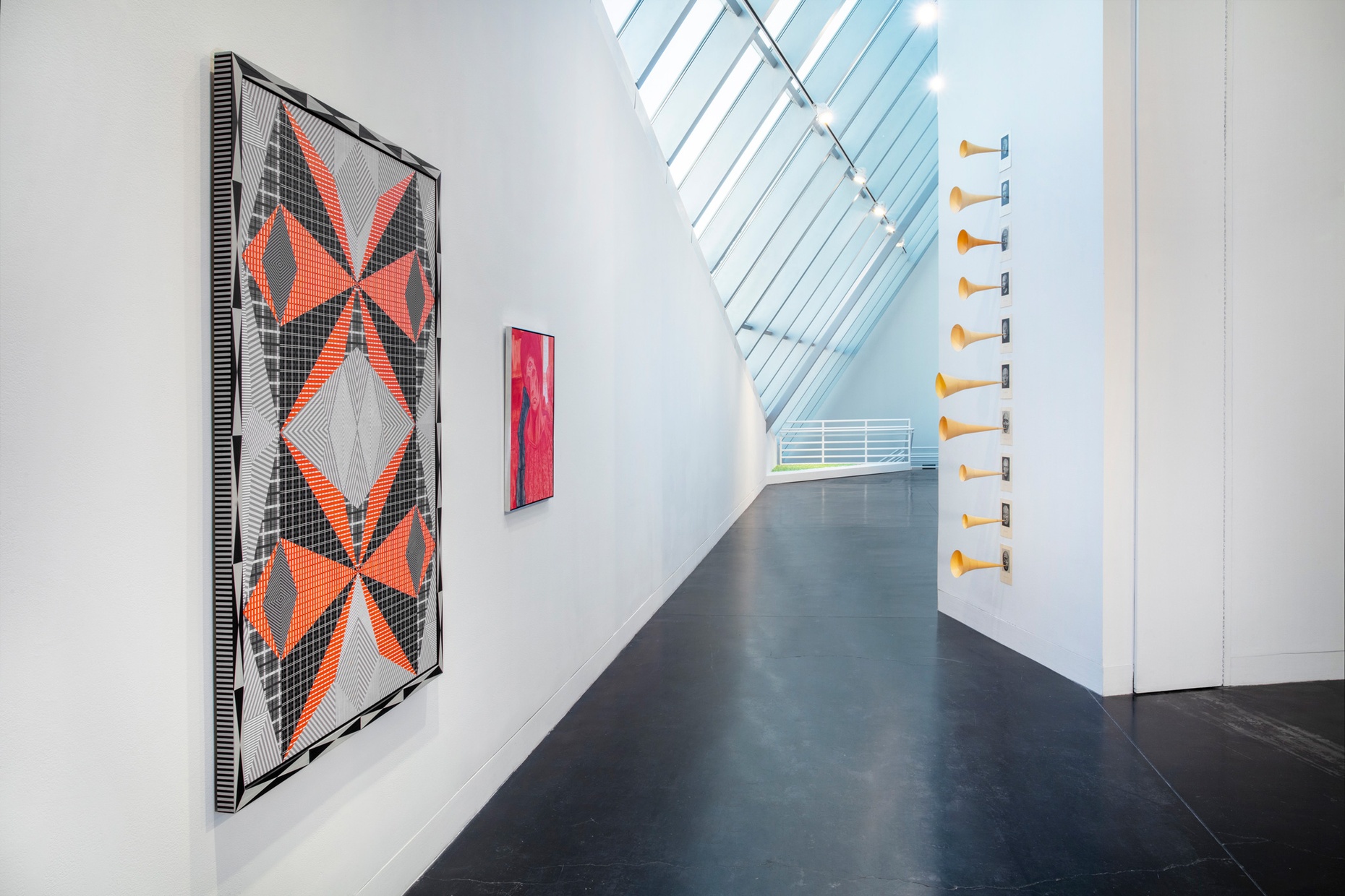 Two colorful, abstract works hang on a white wall in a hallway across from a sculpture of a vertical row of photographs with paper trumpets coming out of each photo.