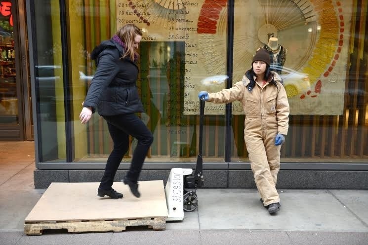 A woman dances on a crate trolley. On the left another woman holds the crate trolley hanlde