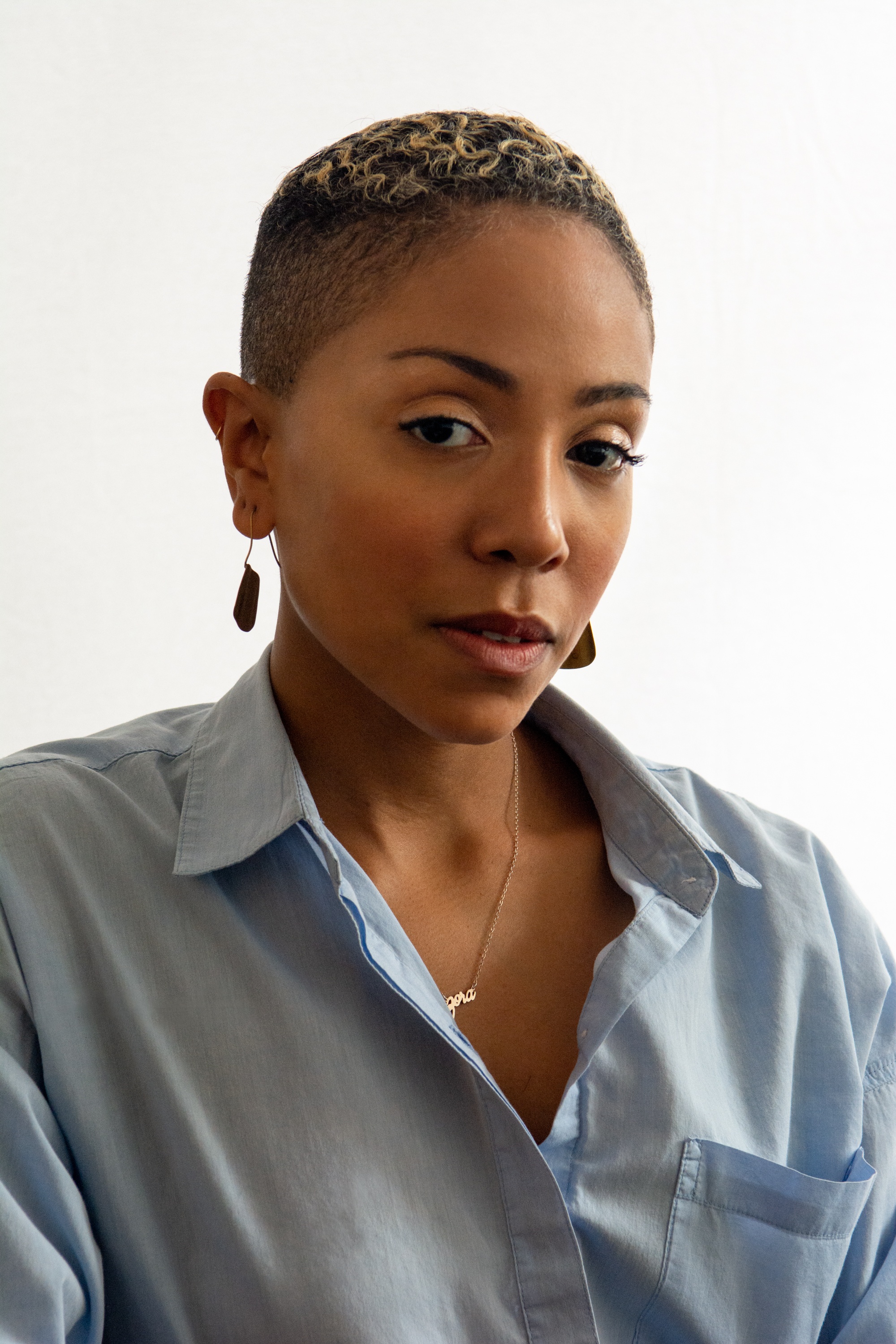 A portrait of Zora Howard with short hair, wearing earrings and a blue collared shirt