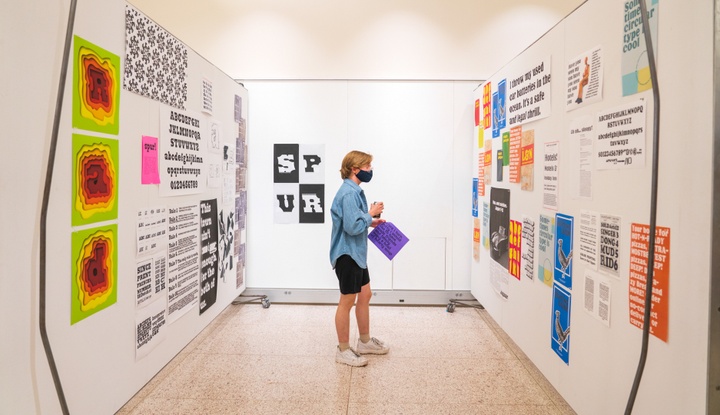Person stands in a gallery space between two walls of typographic work, including type samples, patterns, and singular illuminated letters.