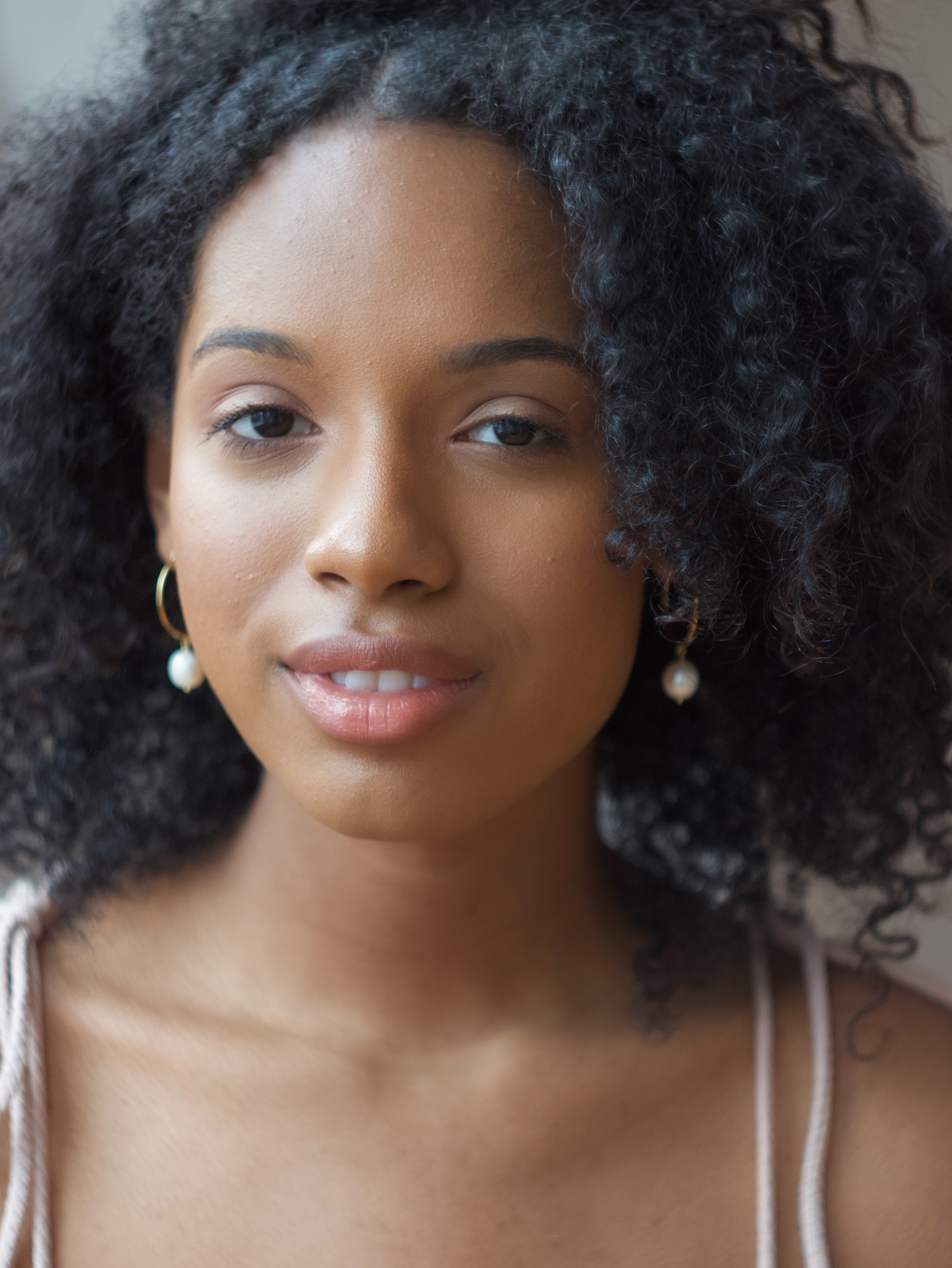 A Black woman with curly shoulder-length hair looking at the camera with one eyebrow raised. She wears small gold hoop earrings with pearls on the ends