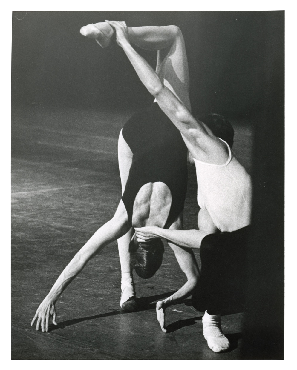 A black and white photograph of two ballet dancers, one balanced on one foot with body upside down and back to the camera, the other provides support at the head and raised foot.