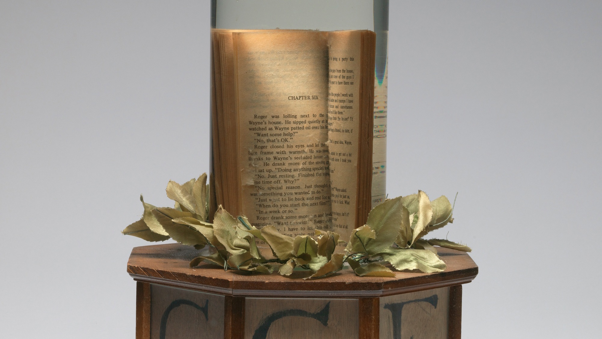 Atop a wooden pedestal is fake greenery at the base of a glass jar containing an open book in water.
