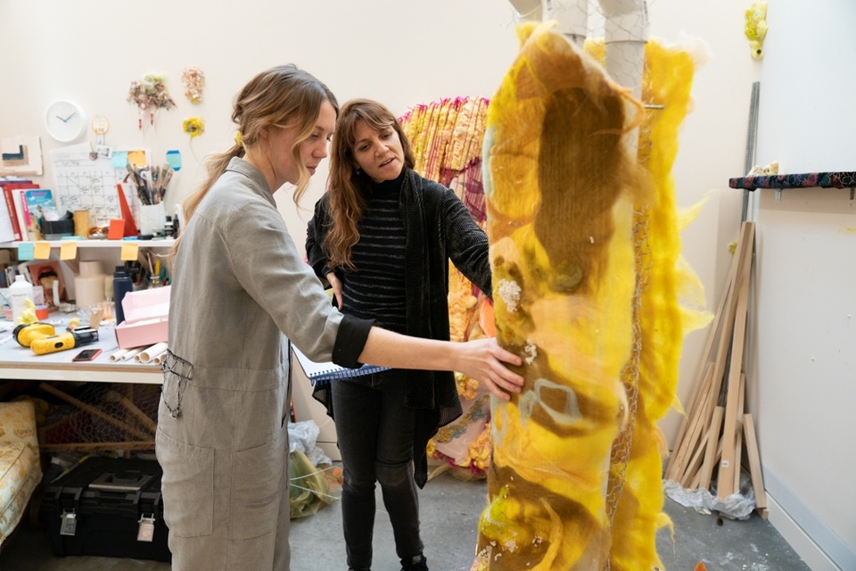 Two people inspect a large-scale felt sculpture in a studio space containing a desk, toolboxes, and a pile of lumber.