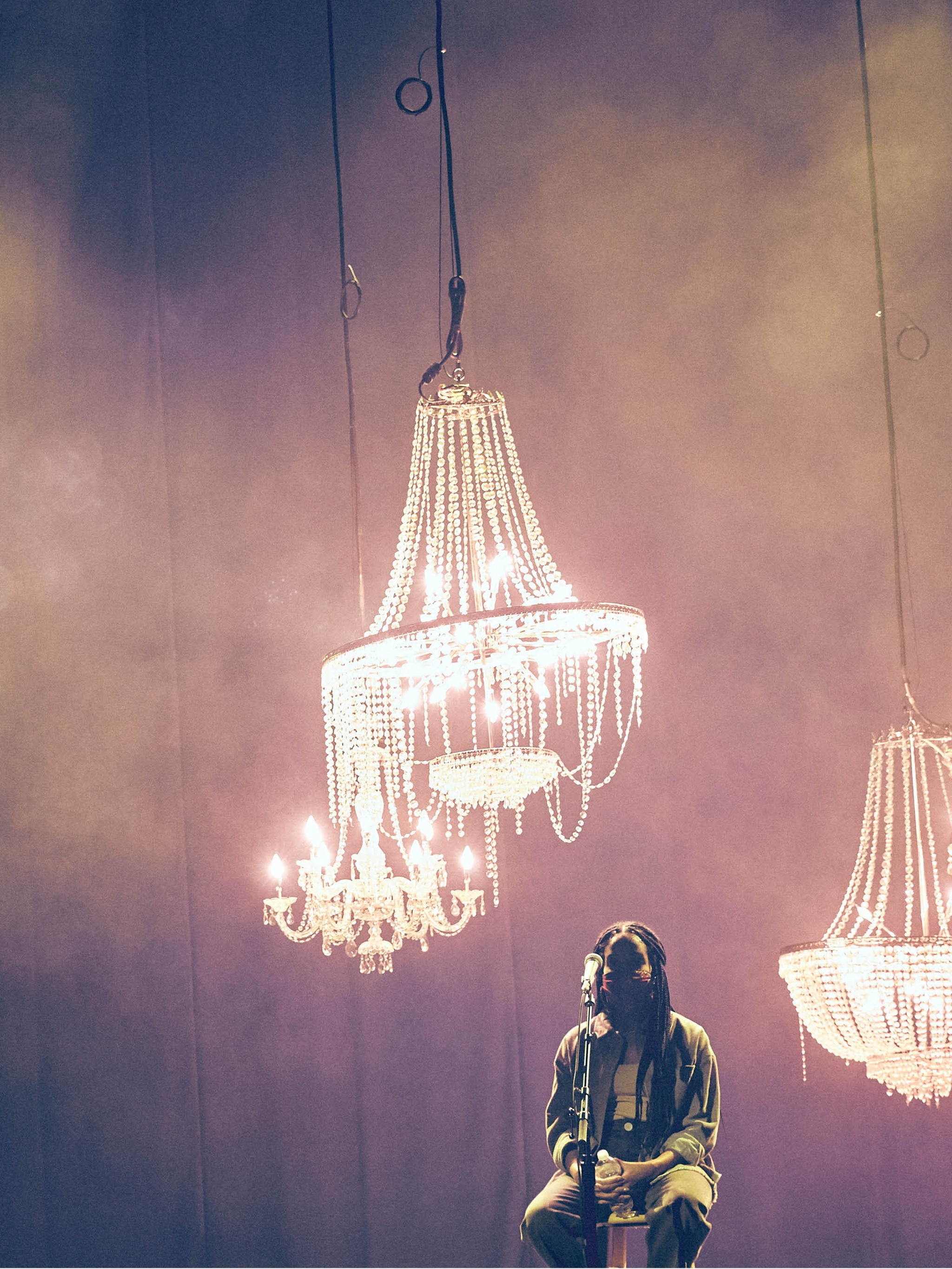 A woman with long braids sitting at a microphone in a hazy space with chandeliers hanging behind her