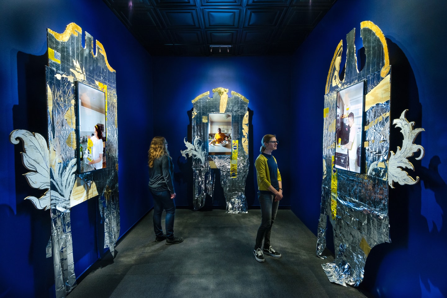 Three channel video installation in a blue room. Each video screen is framed by an ornate structure with silver and yellow marks 