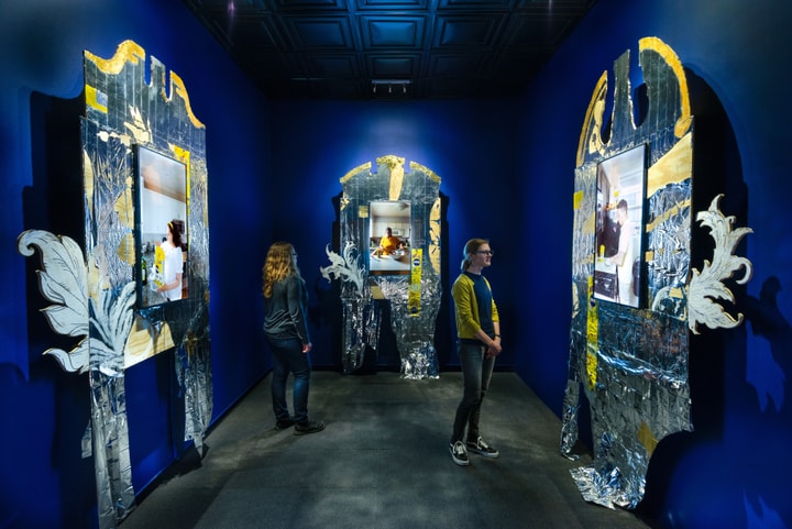 Three channel video installation in a blue room. Each video screen is framed by an ornate structure with silver and yellow marks 