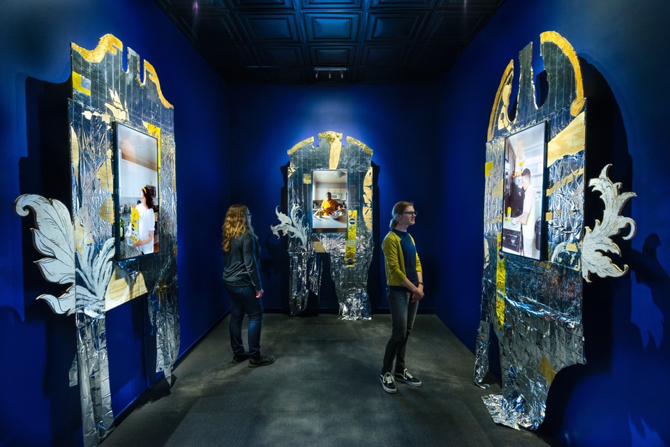 Three channel video installation in a blue room. Each video screen is framed by an ornate structure with silver and yellow marks 