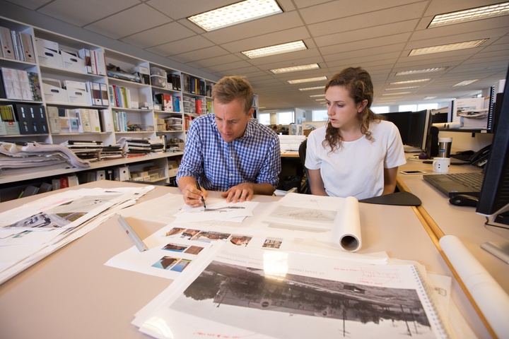 Two people sit on the same side of a table. One draws on a large paper while the other watches.