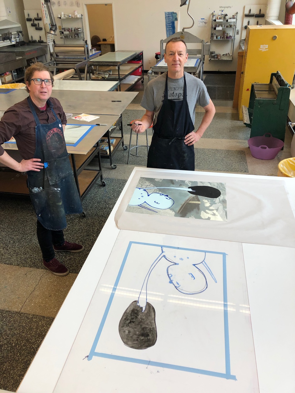 Tom Reed and David Humphrey pose for a photo in front of a table with two large images prepped for print in the printing studio.  