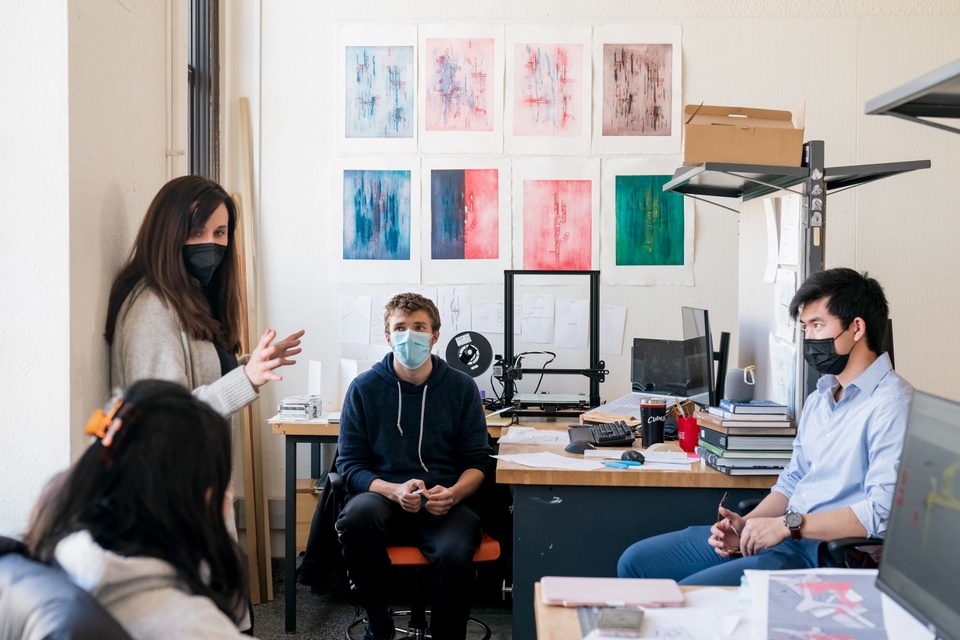 A standing instructor talks to three seated students in an alcove of a studio with colorful relief prints on the wall and a 3D printer sitting on the desk behind them.