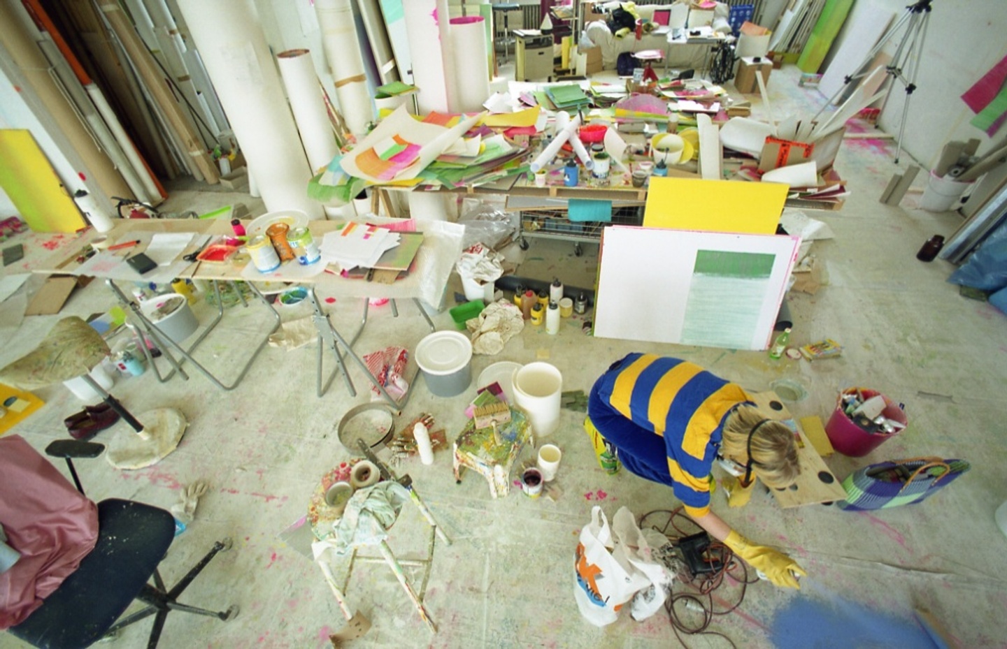 A painter working in a studio, strewn with papers and buckets, spraying blue paint onto the floor