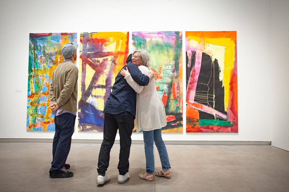 Set of four colorful abstract paintings on vertical canvases. Two gallery visitors embrace in front of them.