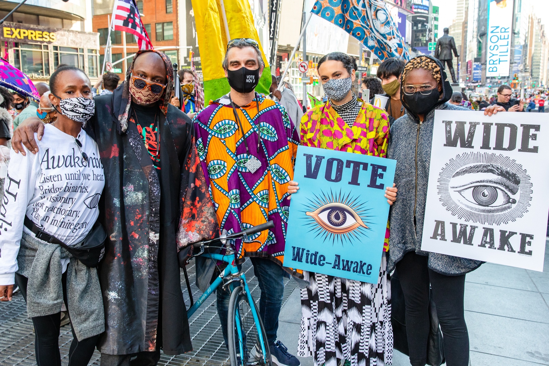 A photograph of five people standing close, wearing masks, a crowd of people behind them. Two people hold posters that say “VOTE Wide-Awake” and “WIDE AWAKE”; both feature the eye.